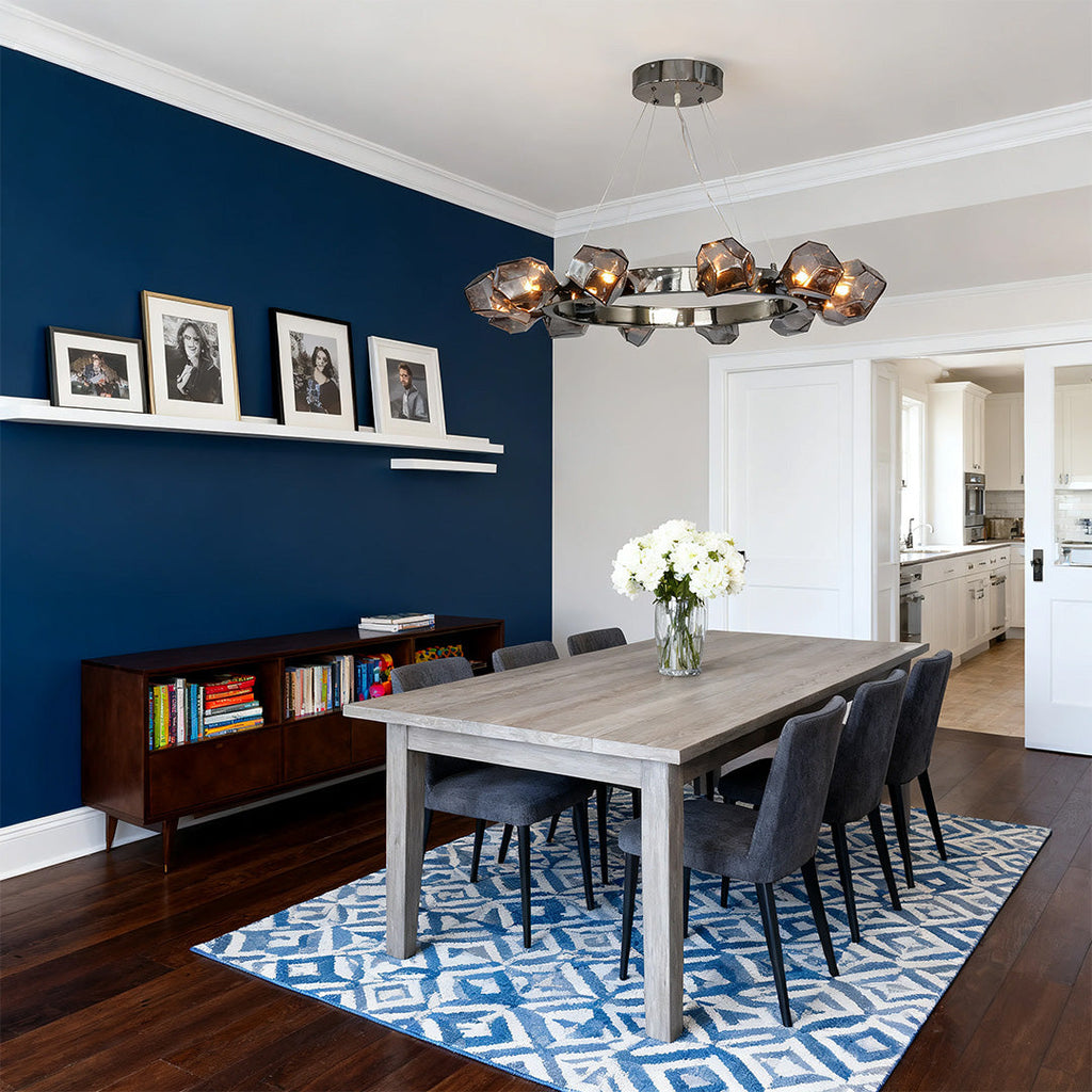 Sleek black round chandelier providing light over a large gray dining table set against a bold blue accent wall and blue patterned rug.
