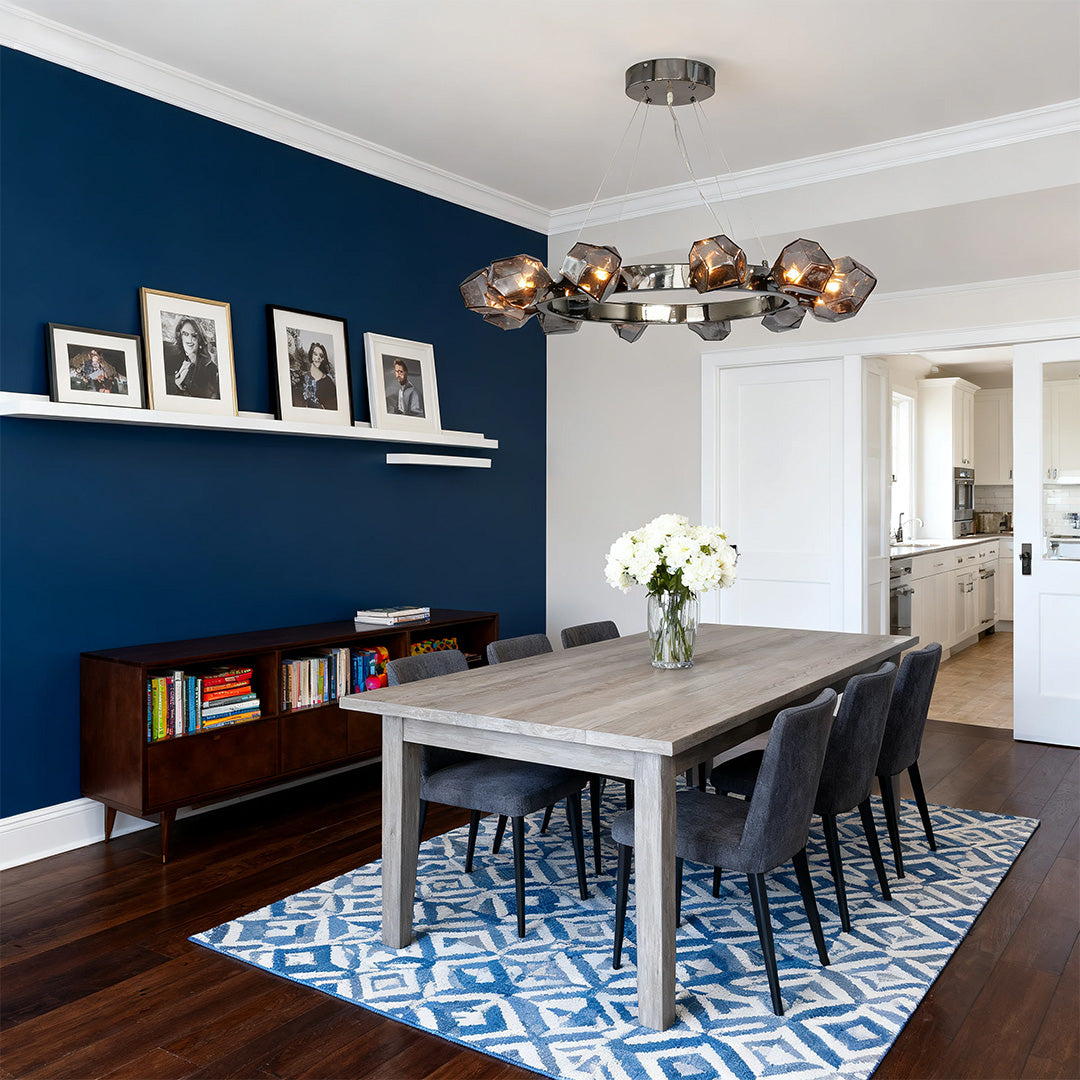 Sleek black round chandelier providing light over a large gray dining table set against a bold blue accent wall and blue patterned rug.